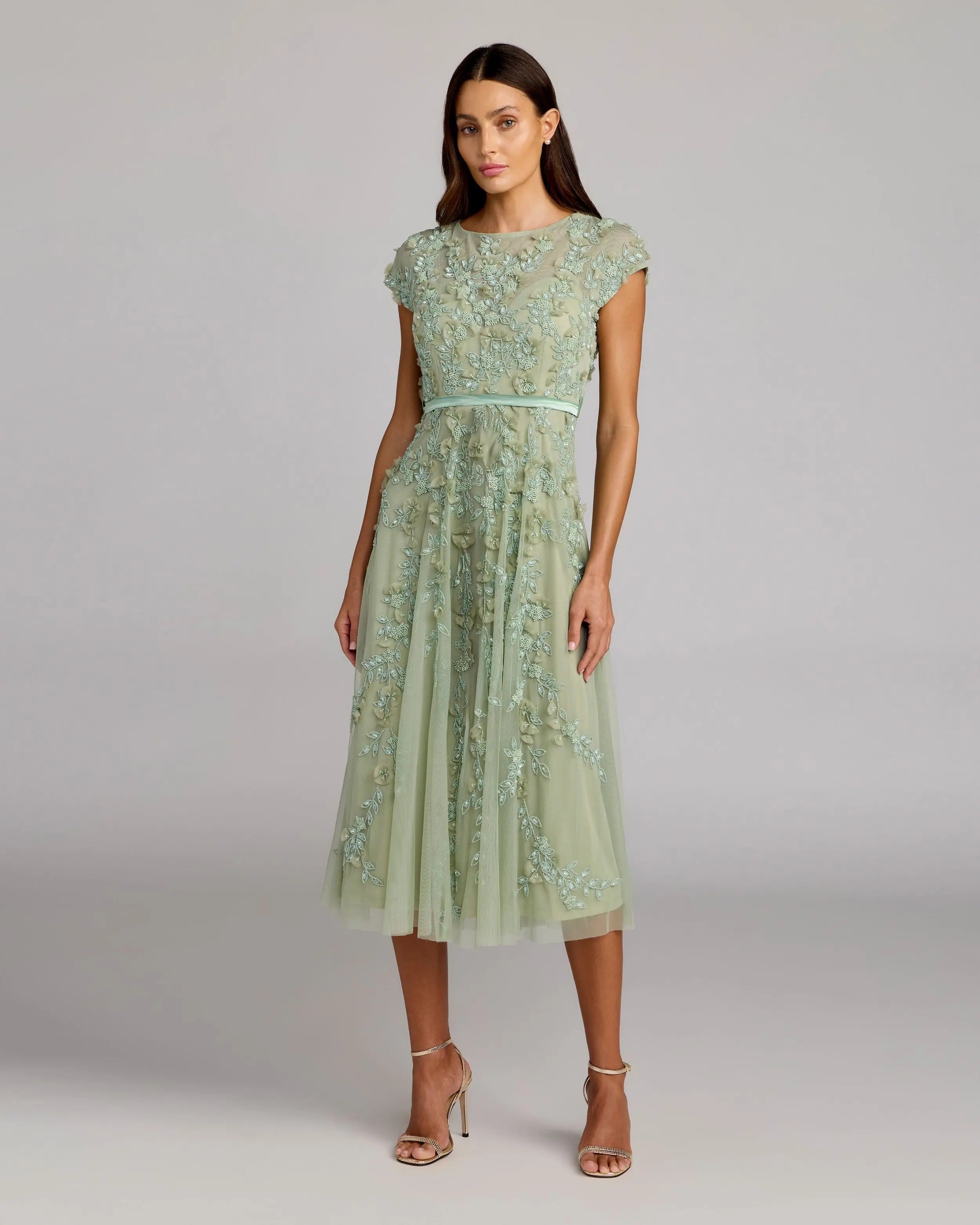 A woman wears the Mac Duggal 94339 midi wedding guest dress in sage, featuring cap sleeves, floral embroidery, a thin belt, and strappy heels—an elegant choice for a wedding reception.